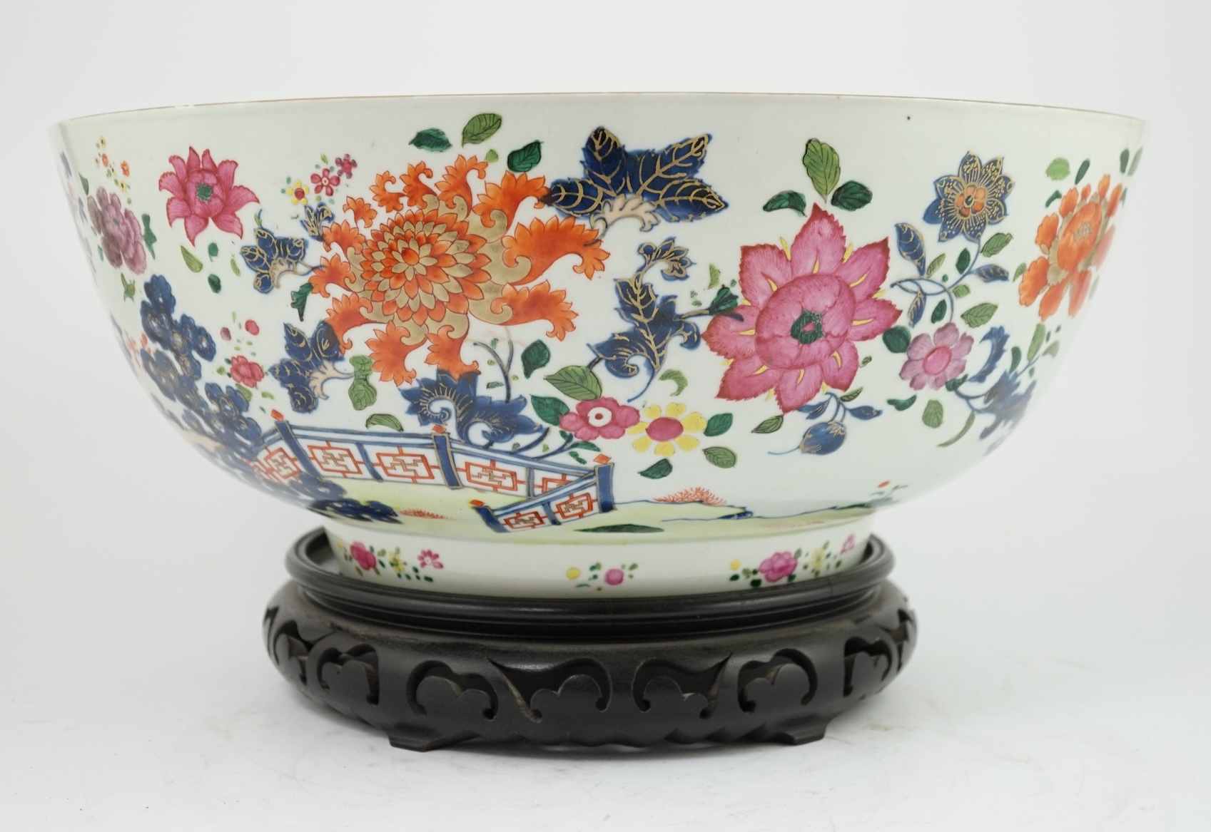 A large Chinese famille rose punch bowl, Qianlong period, the exterior painted with flowers in a fenced garden, the interior with a flower bouquet within a garland of flowers, 40.5cm diameter, wood stand. Condition - som